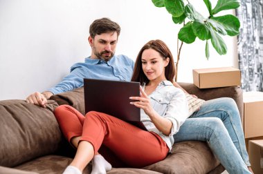 Evdeki rahat oturma odasındaki kanepede oturan genç çift. Erkek dizüstü bilgisayara bakıyor, kadın eşyaları gösteriyor ve alıyor, karton kutuları yeni bir eve götürmek için arama hizmeti veriyor.