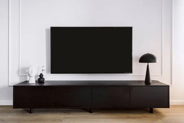 stock image Modern tv screen hanging on white wall in living room with minimalistic interior. Black wooden stand under device. Home decor and lamp on cabinet. Household electronics concept