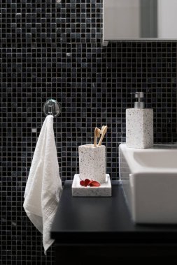 Dikey banyo görüntüsü, tahta diş fırçası sabun şişesinin yanında ve siyah kaplama mozaik duvardaki askıda beyaz, taze ve temiz yüz havlusu.