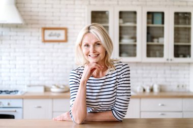 Kameraya bakıp gülümseyen olgun bir kadının portresi. Mutfak ahşap masasında arka planı bulanık oturan bir kadın. Gündelik giysiler içinde büyüleyici bir ev hanımı sabahını evde geçiriyor.