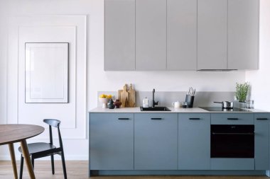 Modern iç ve ev aletlerine sahip mutfak, elektrikli fırın, indüksiyon hob, lavabo su musluğu, mavi dolap, sandalyeli yemek masası ve duvarda boş resim.