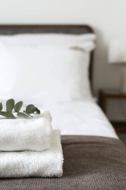 Stack of white bathroom towels with green branch of eucalyptus on top of made bed in guest bedroom. Hospitality service, hotel business and tourist resort concepts. Selective focus on textile clipart