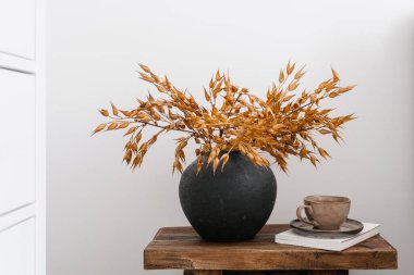 Ahşap tezgahta siyah kil vazoya düşen çiçekler. Kuru bitkilerle dolu bir bank, sabah kahve fincanı ve beyaz fotokopi duvarı arka planına karşı odada kitaplar. Oturma odasının iç kısımlarında hareketsiz yaşam kompozisyonu