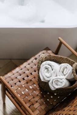 Temiz pamuk havluların yüksek açılı görüntüsü hasır sepete katlanmış. Konforlu banyoda sandalyede tekstil. Banyo köpüklü küvet. Kişisel bakım, hijyen, vücut bakımı ve güzellik kavramları.