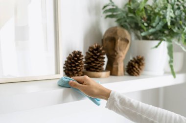 Mavi bez tutan ve evdeki ahşap beyaz rafı temizleyen kadına odaklan. Mobilyaların tozunu dekoratif elementlerle temizliyorum. Ev işi kavramı.