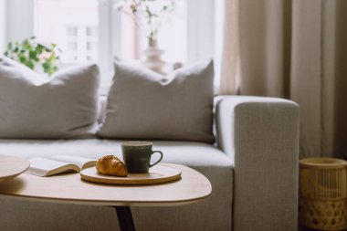 Modern evdeki bulanık oturma odasının iç kısımlarına karşı sehpanın üzerinde seramik bardaklı, sıcak içecek ve taze fırın bulunan bambu tepsisine odaklan. Dinlenme ve kitap okuma zamanı.