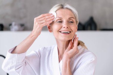 Gülümseyen orta yaşlı kadın arındırıcı tonik kullandıktan sonra sağlıklı ve saf cildinin tadını çıkarıyor. Kadın banyoda pamuk pedlerle yenilenen kozmetik ürünler uyguluyor. Cilt bakımı kavramı