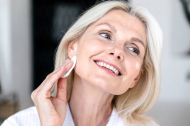 Orta yaşlı bir kadın pamuk yastıkla yüz sulandırıcı tonik uyguluyor, aynaya bakıyor, gülümsüyor ve cilt bakımından zevk alıyor. Kırışıklık önleyici kozmetik ürünü kullanan bir kadın.