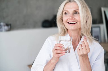 Diyet ve beslenme konsepti. Sağlıklı yiyecekler. Mutlu gülümseyen 50 'li yaşlardaki yaşlı kadın Omega 3 ile kapsül kullanıyor. Bir bardak su tutuyor. C, D, E vitamini, balık yağı..