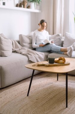 Oturma odasında çay fincanı ve kruvasan olan ahşap sehpaya odaklan. Kaygısız bir kadının kağıt kitap okuması, hafta sonunun keyfini çıkarması...