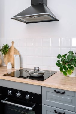 Vertical shot of kitchen interior. Frying pan with lid on induction hob, integrated electric oven, cabinet with wooden countertop, modern extractor hood, tiled wall, cutting board and green houseplant clipart