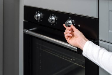 Modern mutfaktaki yeni siyah temiz elektrikli fırının regülatör düğmesini evde yapılmış ekipmanlarla kadın elinin açması üzerine seçici bir odaklanma. Yemek pişirme süreci