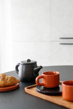 Seramik çaydanlık, demlenen bitkisel çay, ahşap tepside fincanlar ve taze Fransız kruvasanı mutfak masasında, beyaz arka planda fotokopi alanı ile birlikte. Kahvaltı konsepti