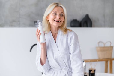 Bornoz giymiş orta yaşlı bir kadın banyoda duş jeli olan bir şişe tutuyor. Cilt bakımı için profesyonel kozmetik ürünler kullanan gülümseyen bir kadın. Güzellik tedavi konsepti