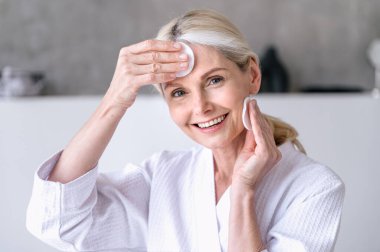 Cilt bakımı. 50 yaşında, gülümseyen, neşeli bir kadın banyoda pamuk pedlerle makyaj yapıyor. Olgun cilt için kozmetik sütle yüz temizleme.