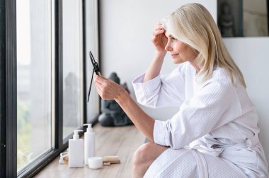 Beyaz bornozlu orta yaşlı bir kadının yaşlanmayı önleyen kozmetik ürünler kullandıktan sonra yüzüne ve aynaya bakıp cildine baktığı profil görüntüsü. Cilt bakımı ve kendine bakım konsepti.