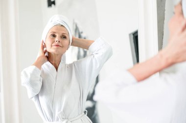Aynanın önünde duran, duştan sonra bornoz ve havlu giyen olgun bir kadın yansımaya bak, banyoda sabahtan zevk al. Bakım, cilt bakımı, saç bakımı prosedürü