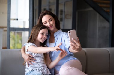 Sevimli kızıyla kucaklaşan neşeli anne kadın, akıllı telefon kamerasına bakıyor ve evdeki kanepede otururken selfie çekiyor. Mutlu aile eğlenceleri birlikte vakit geçirin.. 