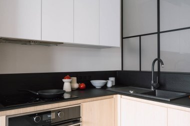 Cihazları, elektrikli fırın kaputu, fırını ve lavabosu olan modern bir mutfak. Beyaz dolapların altındaki ev dekoruna yakın siyah mermer tezgahta cam seramik yüzey üzerinde kızartma tavası.