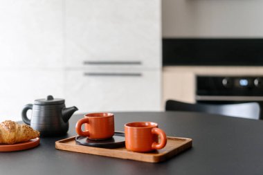 Bambu tahtasına seramik bardaklar, çaydanlık ve yemek odasındaki fırın ile mutfak içi bulanık tabaklar üzerine seçici bir odak noktası. Evde kahvaltı.