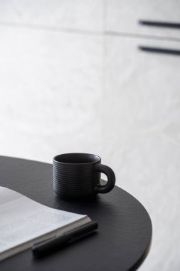 Açık dergi, kağıt kitap ya da günlük dergisi yakınındaki ahşap masada sabah kahvesi olan siyah seramik fincanı kapat. Seçici odak, natürmort nesneler, alan arkaplanı kopyala