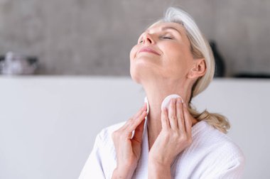 Sağlıklı pürüzsüz cilt için pamuklu sünger kullanan olgun bir kadın. Kadın makyajı silinmiş ve ev banyosunda günlük cilt bakımının keyfini çıkarıyor..