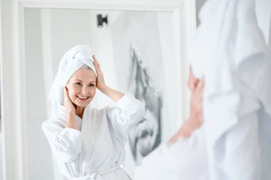 Banyo havlusuna sarılı mutlu kadın, yansımasına bak, duştan sonra sabah rutininin tadını çıkar. Kadın banyoda duruyor. Hijyen, spa prosedürü, güzellik tedavi kavramları