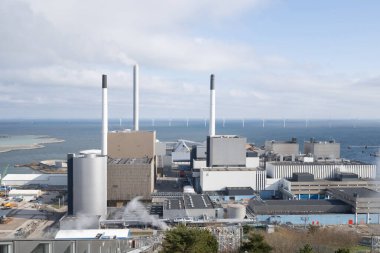 Sahilde büyük bir endüstriyel kompleks. Uzun bacaları ve uzak mesafede rüzgar türbinleri manzarası var.