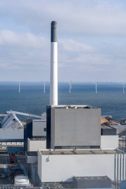 Yüksek bacalı büyük bir endüstriyel kompleksin dikey görüntüsü arka planda birkaç açık deniz rüzgar türbini ile bir rıhtımda duruyor.