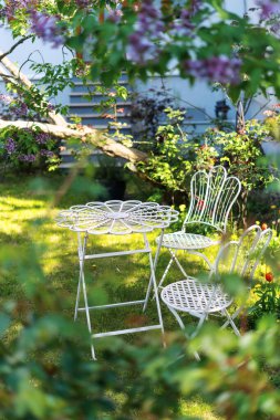 Beyaz bir teras masası ve sandalyeler yemyeşil bir çimenlikte, yemyeşil yapraklar ve çiçekli bitkilerle çevrili. Rahat dinlenmek için bahçe mobilyaları ve evin dışında boş zaman geçirmek için.