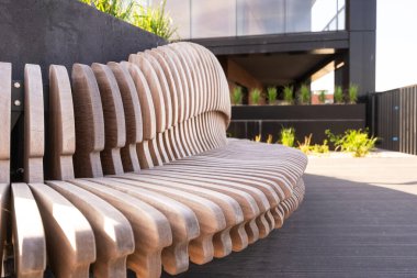 Modern, kıvrımlı bir ahşap bank şehir parkında oturur, arka plan ise siyah yüzlü modern bir binadır..