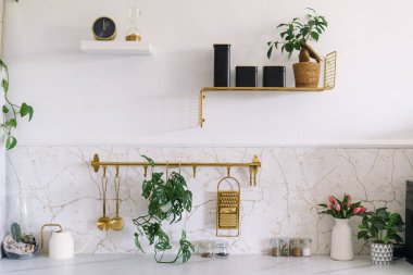 Parlak bir mutfak, mermer bir tezgah, altın raflar, saksı bitkileri, ev dekorasyonu ve şık bir depo sergiler, odada dingin ve modern bir ortam yaratır.