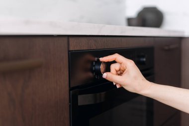 Kırpılmış kadın dönüş düğmesi mutfaktaki fırının sıcaklığını düzenler. Ev hanımı zamanlayıcı ayarlıyor ya da mikrodalgada çözme programını seçiyor. Ahşap dolapta ev aletleri var.