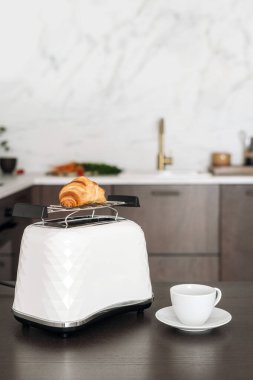 Beyaz modern tost makinesinin yanında duran seramik espresso bardağının dikey görüntüsü taze kruvasanı ısıtıyor. Hamur işi ve sabah kahvesi ahşap mutfak masasının üstünde. İçi bulanık.