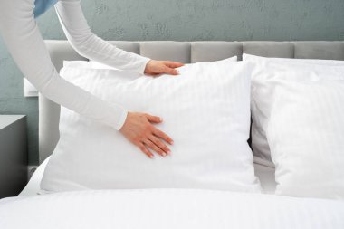 Kadın yatak yapıyor, yeni yastık kılıflarındaki beyaz köpük yastıkları düzleştiriyor. Rahat bir şekilde rahat bir odada, yumuşak bir yatakta uyuyorsun. Otel odası, temizlik servisi.