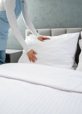 Mavi önlüklü hizmetçi otel odasında yatak yapıyor, yastığı beyaz ve temiz yastık kılıfıyla düzleştiriyor ve çarşafları örtüyor. Ev işleri, hizmetçi kadın için günlük rutin, dikey çekim.