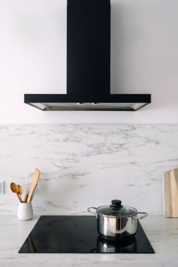 Modern cam seramik fırının üzerine siyah kapüşonlu, paslanmaz çelik tavayla dikey çekim. Mutfağın içinde elektrikli tencere ocağı var. Mermer döşeme, beyaz kopya alanı duvarı