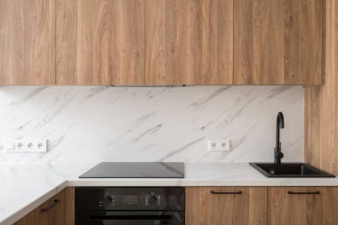 Siyah lavabosu, musluğu ve ocağı olan modern bir mutfak tezgahı. Ahşap dolaplarda yapılmış fırının üzerine indüksiyon hobini. Mutfakta fayanslı duvara dayalı ev aletleri.