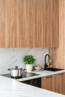 Modern iç mekan elementi. Siyah bir sobanın üzerinde paslanmaz çelik tencere. Beyaz tezgahlı, mermer kaplama duvarlı ve ahşap dolaplı mutfağın yan manzarası. Lavabonun yanında küçük bir bitki ve mutfak malzemeleri var.