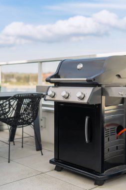 Siyah gaz ızgarasının dikey görüntüsü teras sandalyesi ve arka planda masası olan bir teras avlusunda duruyor. Gökyüzünün altında mavi ve beyaz bulutlu barbekü ekipmanları. Hafta sonları dışarıda yemek pişirmek.