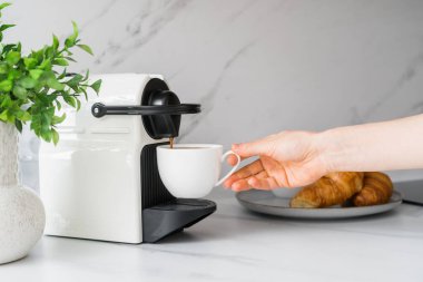Espresso makinesi kapuçinoyu fincana doldurur. Kadın sabah kahvesini evde yapıyor. Mutfak tezgahındaki kahve makinesi mermer arka planda hamur işine yakın.
