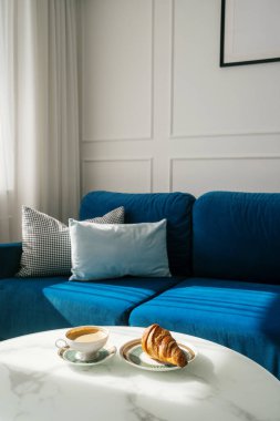 Bir fincan kahve ve oturma odasındaki beyaz mermer kahve masasında bir tabakta kruvasanla dikey çekim. Espresso ve Fransız pastası, yastıklı mavi bir kanepenin önünde. Evde sabahlar