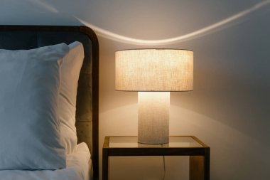 Yatak odasının iç kısmının dikey görüntüsü, yastıklı çift kişilik yatağın yanındaki komodinin üzerinde modern okuma lambası. Konforlu dairede akşam ışığı. Rahat otel odası, yıldırım teçhizatı