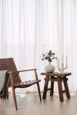 Yumuşak ekoseli retro sandalye, beyaz seramik vazoda okaliptüs ve arkadaki pencere tülüne karşı evdeki ahşap sehpanın üzerinde mum tutacağı tasarla. Tarafsız oturma odası iç mekan kavramı