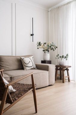 Modüler kanepeli yaşam alanı, ahşap mobilyalı ev dekorasyonu ve rahat sandalyeli. Penceresinde hafif tüller olan ferah, aydınlık bir oda. Koltuk ve kanepe, sehpanın yanındaki çiçeklere yakın.