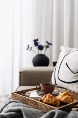 Arka planda bulanık odaya karşı taze kruvasan ve siyah sabah kahvesi ile kahvaltı tepsisine odaklan. Bir fincan bitki çayı ve tatlı hamur işi ile kanepe. Otel servisi, hafta sonu evde.