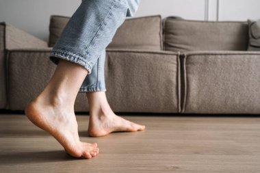 Yeraltı ısıtması ve konforlu ev kavramları. Kadın oturma odasındaki ahşap zeminde yalınayak yürüyor, kapat. Dişi ayaklar dairede doğal parke tahtasına basıyor, seçici bir şekilde yaya olarak.