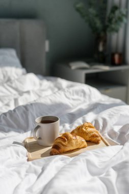Bir fincan sabah kahvesi ve taze pişmiş kruvasanlar ahşap bir tepsiye konur ve yatak odasında rahat bir kahvaltı ortamı yaratır. Lüks otelde oda servisi, evde sakin bir hafta sonu.