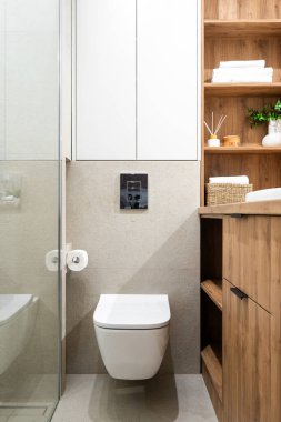 Duvara monte edilmiş beyaz bir klozet, kağıt tutacağı ve cam duş kapısına yakın bir dolabı olan modern bir banyo.