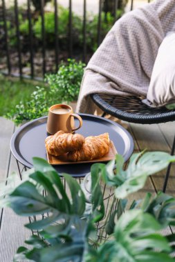 Sıcak balkon kahvaltısı, taze kruvasan ve terasta kahve fincanı, dikey çekim. Verandanın yakınındaki küçük masada fincanda Fransız pastasının ve sıcak içeceğin seçici odak noktası.